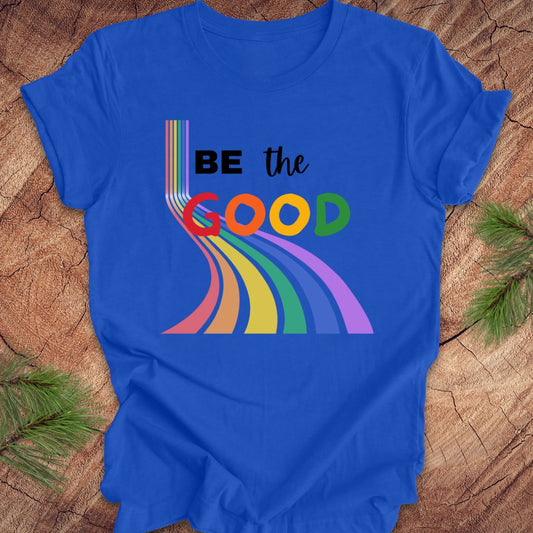 Royal blue t-shirt with rainbow graphic and text, 'Be the Good' on a wood background.