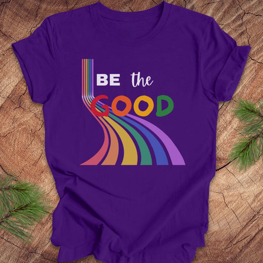 Purple t-shirt with rainbow graphic and text, 'Be the Good' on a wood background.