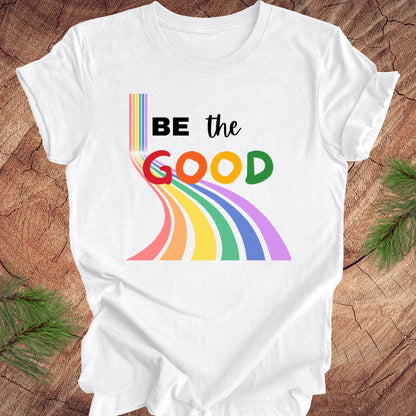 White t-shirt with rainbow graphic and text, 'Be the Good' on a wood background.