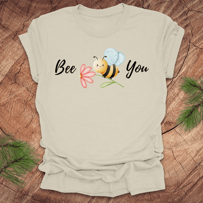 Beige tshirt with a cute bee and the text, "bee you" on a wooden background.