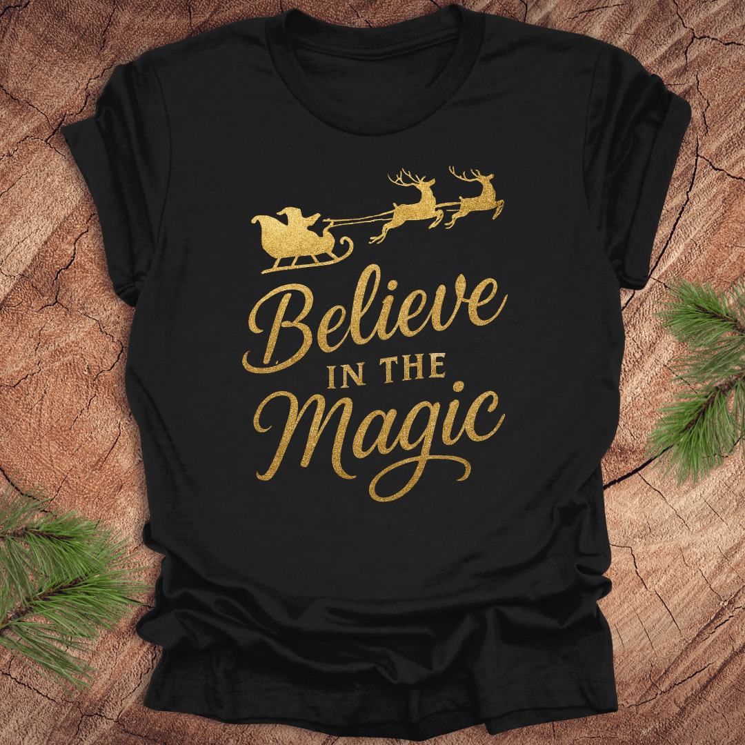 Black t-shirt with gold 'Believe in the Magic' text and Santa sleigh design on a wooden surface.