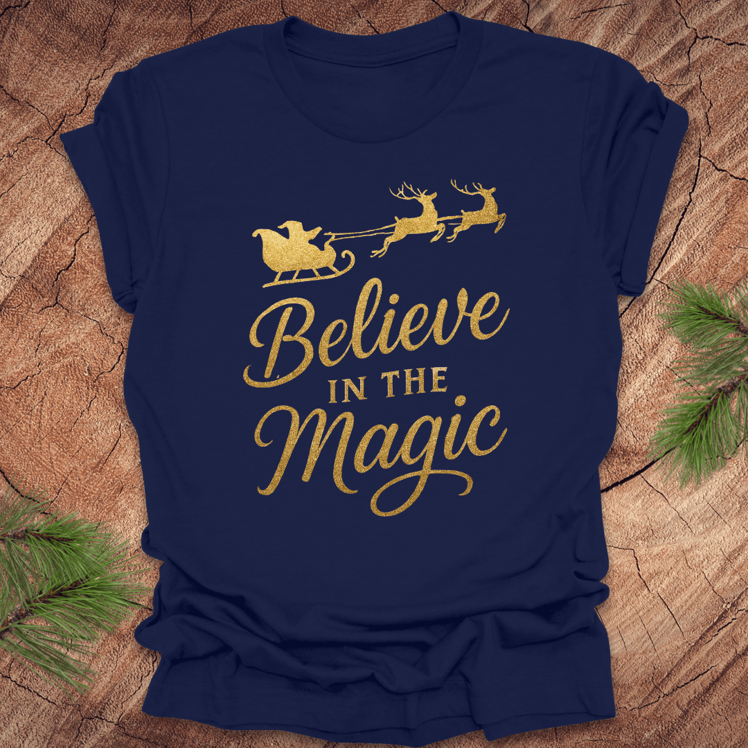 Navy blue t-shirt with gold 'Believe in the Magic' text and Santa sleigh design on a wooden surface.