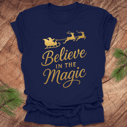 Navy blue t-shirt with gold 'Believe in the Magic' text and Santa sleigh design on a wooden surface.