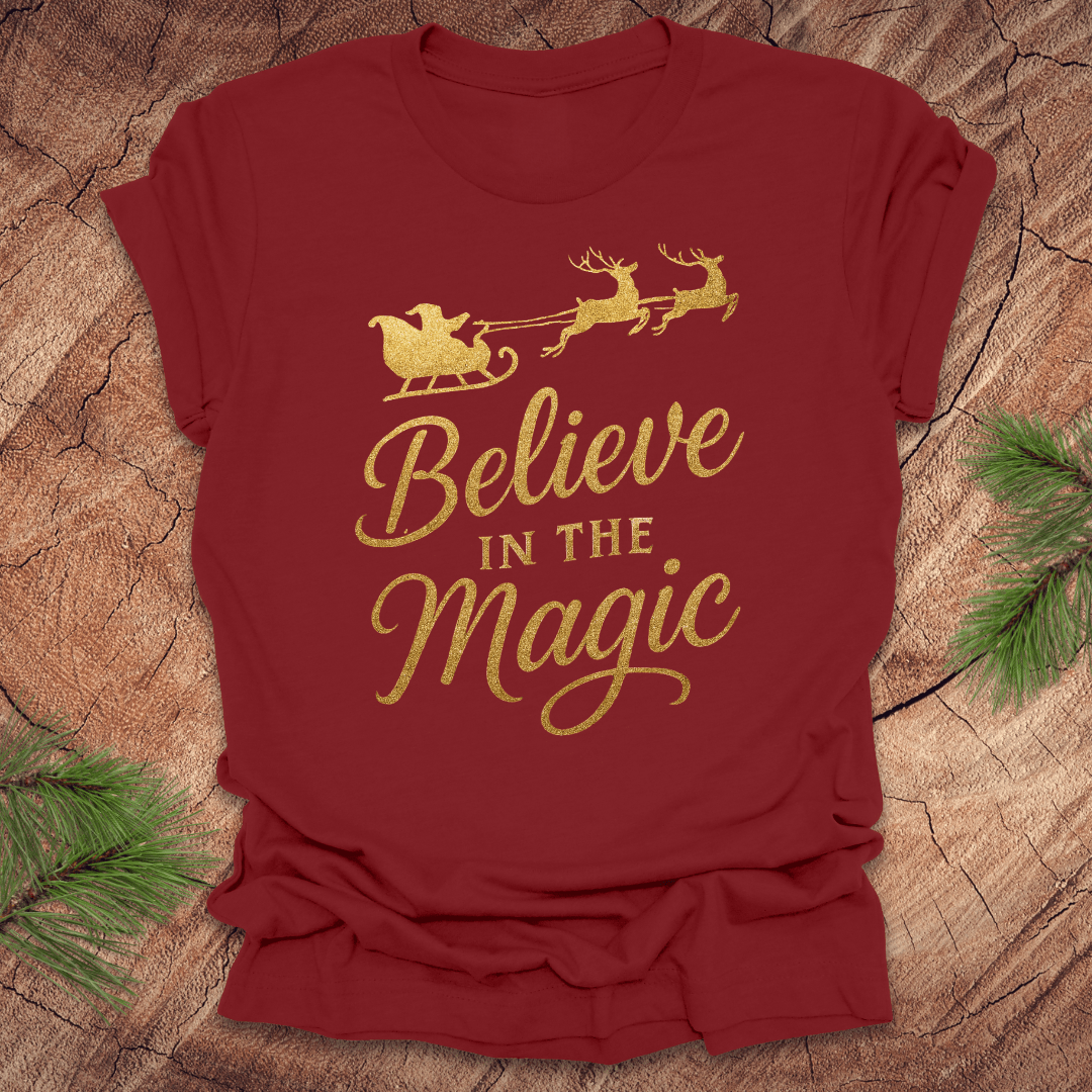 Red t-shirt with gold 'Believe in the Magic' text and Santa sleigh design on a wooden surface.