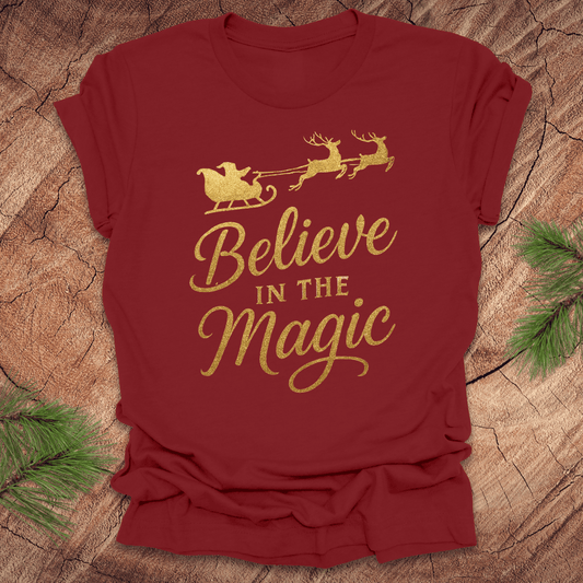 Red t-shirt with gold 'Believe in the Magic' text and Santa sleigh design on a wooden surface.