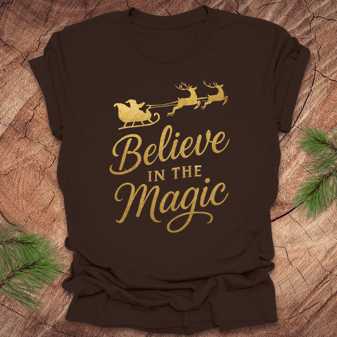 Brown t-shirt with gold 'Believe in the Magic' text and reindeer design on a wooden surface.