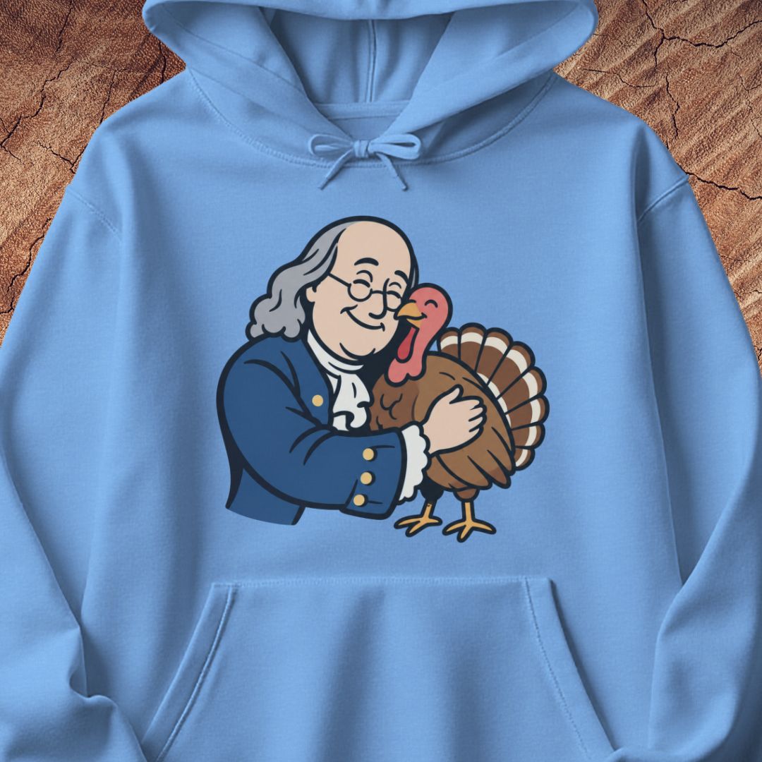A light blue hoodie laid flat on a rustic wood background, featuring a cartoon graphic of Benjamin Franklin hugging a turkey on the front.