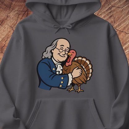 A dark gray hoodie featuring a cartoon graphic of Benjamin Franklin hugging a turkey, displayed against a rustic wood background.