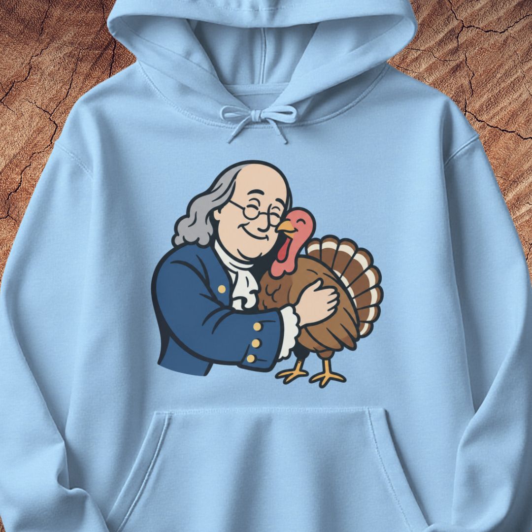 A light blue hoodie laid flat on a textured brown wood background, featuring a cartoon graphic of Benjamin Franklin hugging a turkey on the chest.