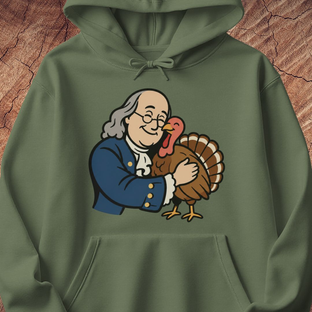An olive green pullover hoodie featuring a cartoon graphic of Benjamin Franklin hugging a turkey on the chest, set against a textured brown wood background.