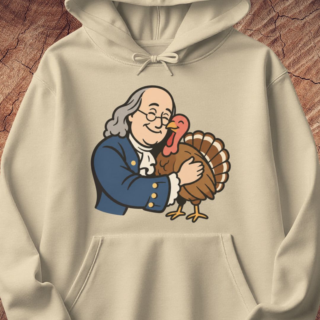 A cream-colored hoodie featuring a cartoon graphic of Benjamin Franklin hugging a turkey, displayed against a textured brown wood background.