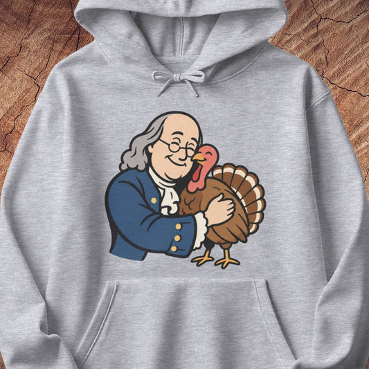 A light heather gray hoodie featuring a cartoon graphic of Benjamin Franklin hugging a Thanksgiving turkey, displayed on a rustic wood background.