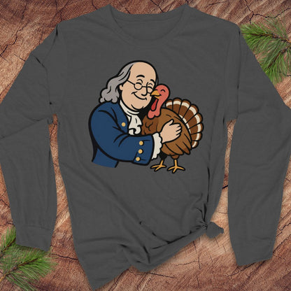 A dark gray long-sleeve t-shirt with a cartoon graphic of Benjamin Franklin hugging a turkey, displayed flat on a rustic wood background with pine needles.