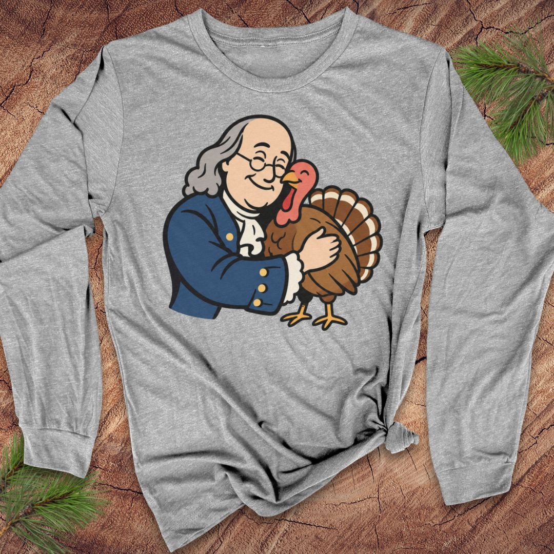 A heather gray long-sleeve t-shirt with a cartoon graphic of Benjamin Franklin hugging a turkey, laid flat on a rustic wood background with pine needles.