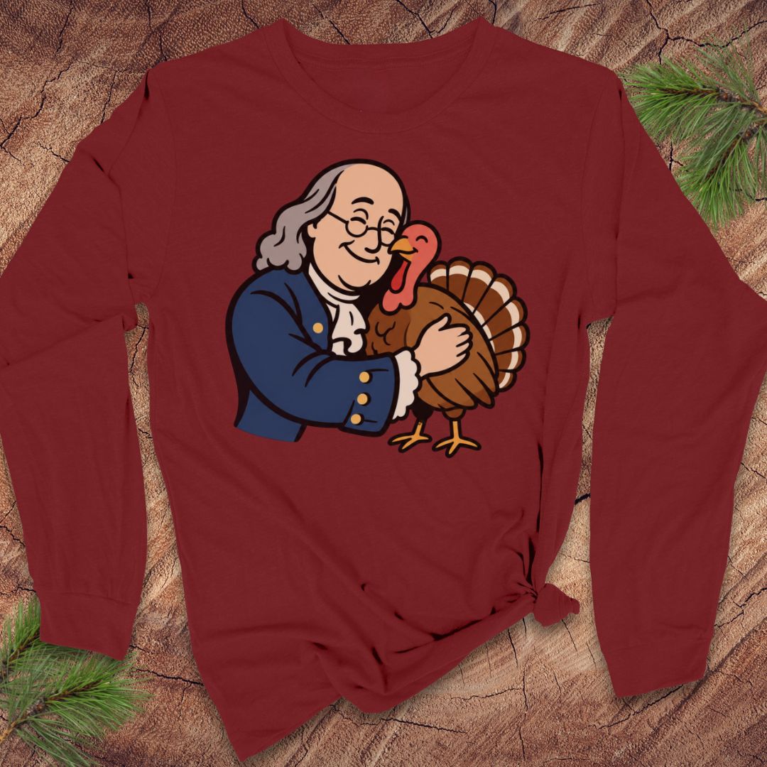 A long-sleeve maroon crewneck t-shirt featuring a cartoon graphic of Benjamin Franklin hugging a turkey, styled flat on a rustic wood background with pine branches.