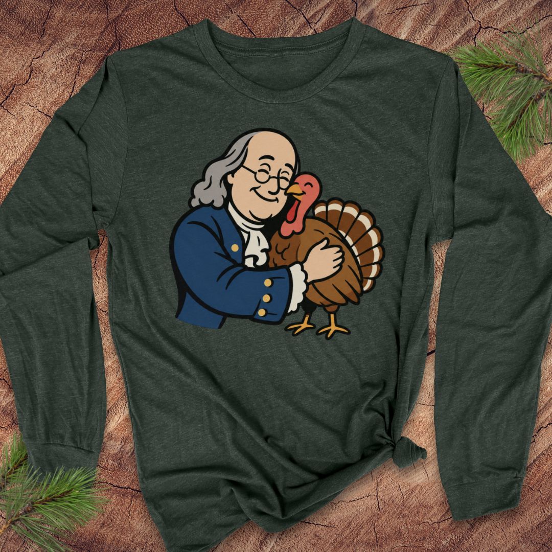 A dark forest green long-sleeve t-shirt with a cartoon graphic of Benjamin Franklin hugging a turkey, laid flat on a rustic wood background with pine needles.