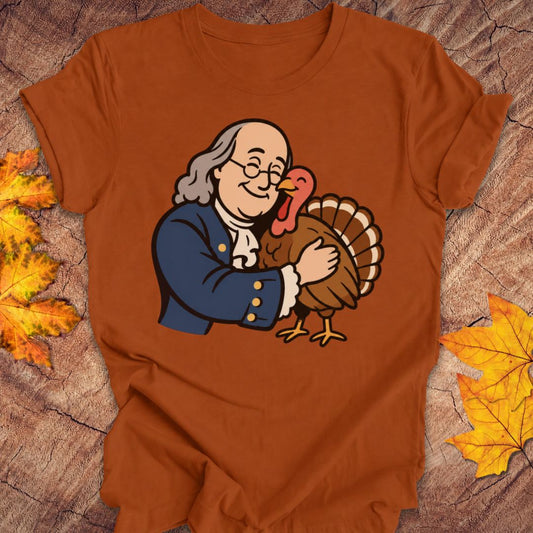 A rust-orange crewneck t-shirt laid flat on a textured wood background with autumn leaves. The shirt features a cartoon graphic of Benjamin Franklin hugging a Thanksgiving turkey.