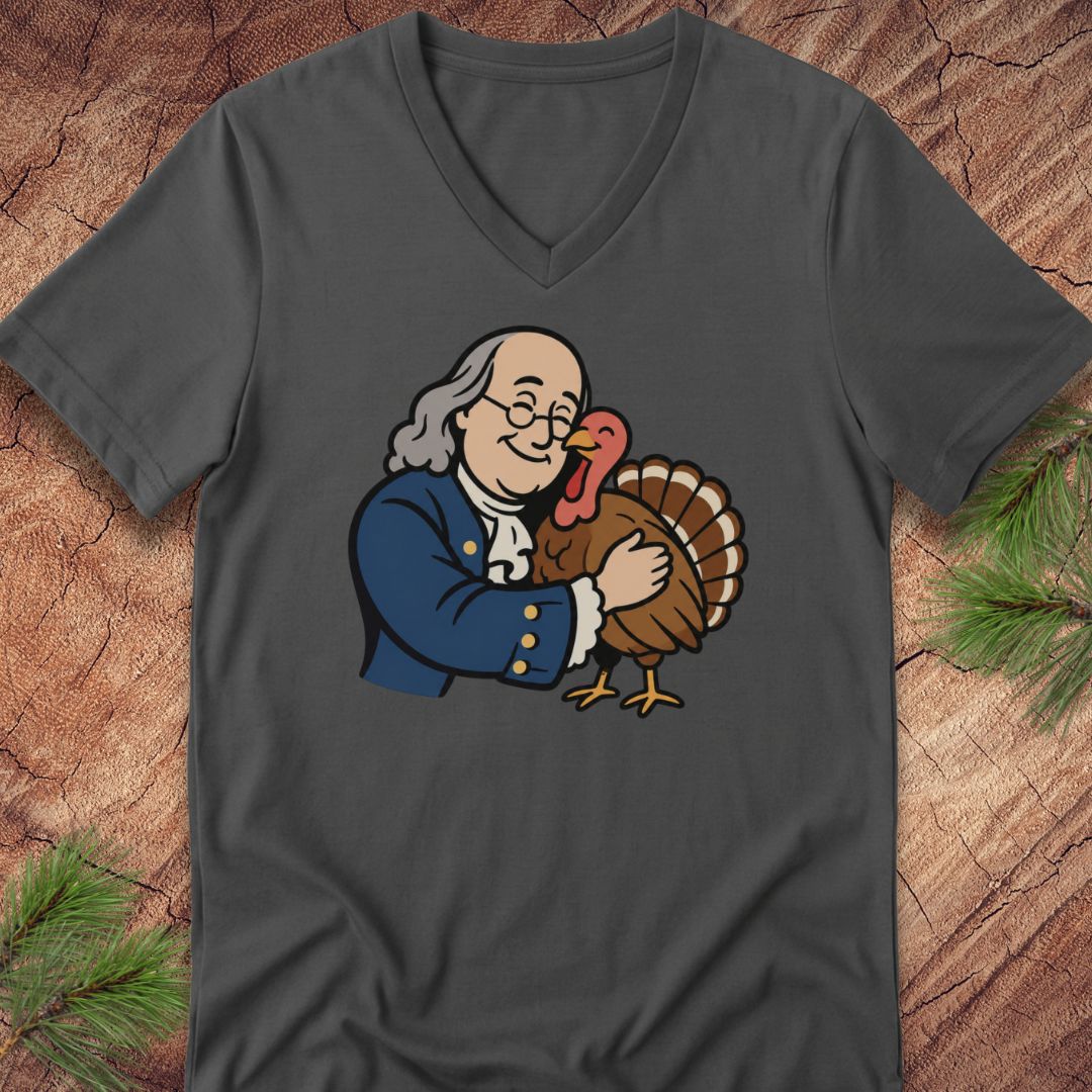 A dark gray v-neck t-shirt featuring a cartoon graphic of Benjamin Franklin hugging a turkey, displayed flat on a rustic wood background with pine needles.