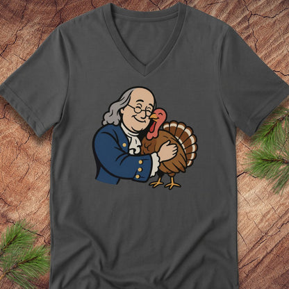 A dark gray v-neck t-shirt featuring a cartoon graphic of Benjamin Franklin hugging a turkey, displayed flat on a rustic wood background with pine needles.