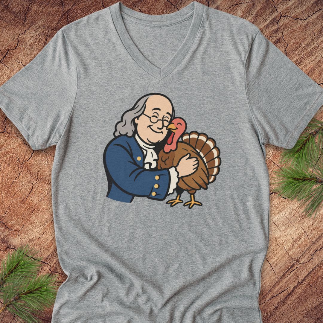 A heather gray V-neck t-shirt laid flat on a rustic wood background with pine needles. The shirt features a cartoon graphic of Benjamin Franklin hugging a Thanksgiving turkey.