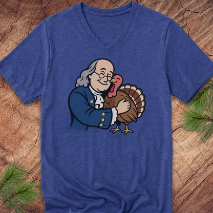 A heathered blue V-neck t-shirt featuring a cartoon graphic of Benjamin Franklin hugging a Thanksgiving turkey, displayed flat on a rustic wood background with pine needles.