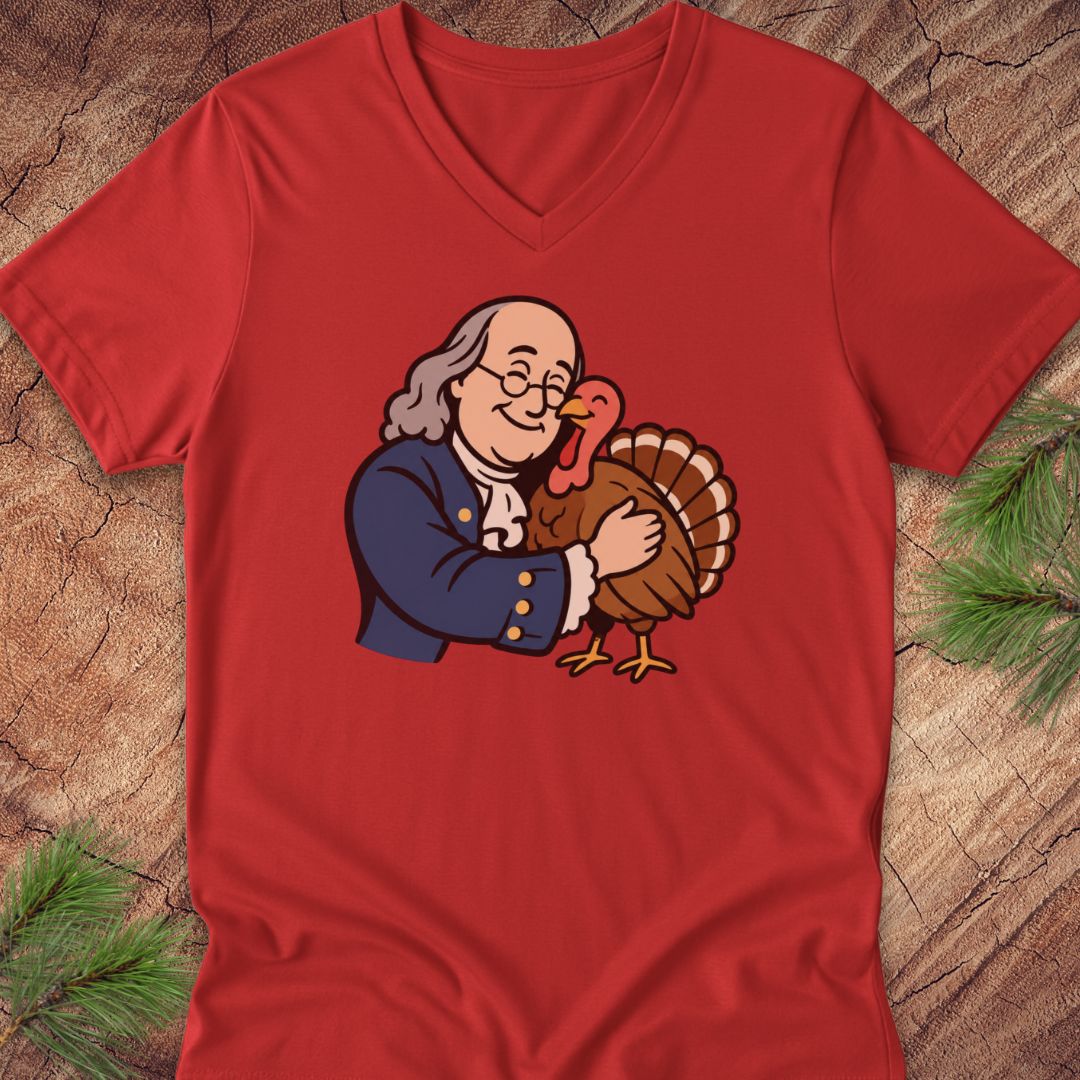 A red V-neck t-shirt laid flat on a rustic wood background with pine needles. The shirt features a cartoon graphic of Benjamin Franklin hugging a Thanksgiving turkey.
