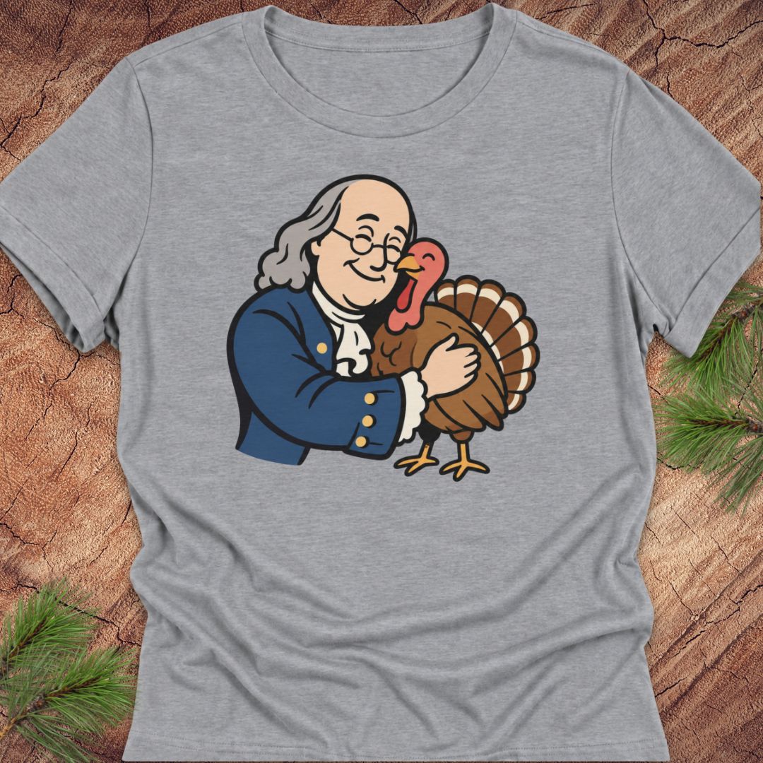 A heather gray short-sleeve t-shirt featuring a cartoon graphic of Benjamin Franklin hugging a turkey, displayed flat on a rustic wood background with pine needles.