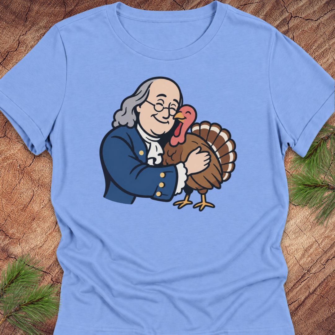 A light blue short-sleeve crewneck t-shirt laid flat on a rustic wood background with pine needles. The shirt features a cartoon graphic of George Washington hugging a brown turkey.