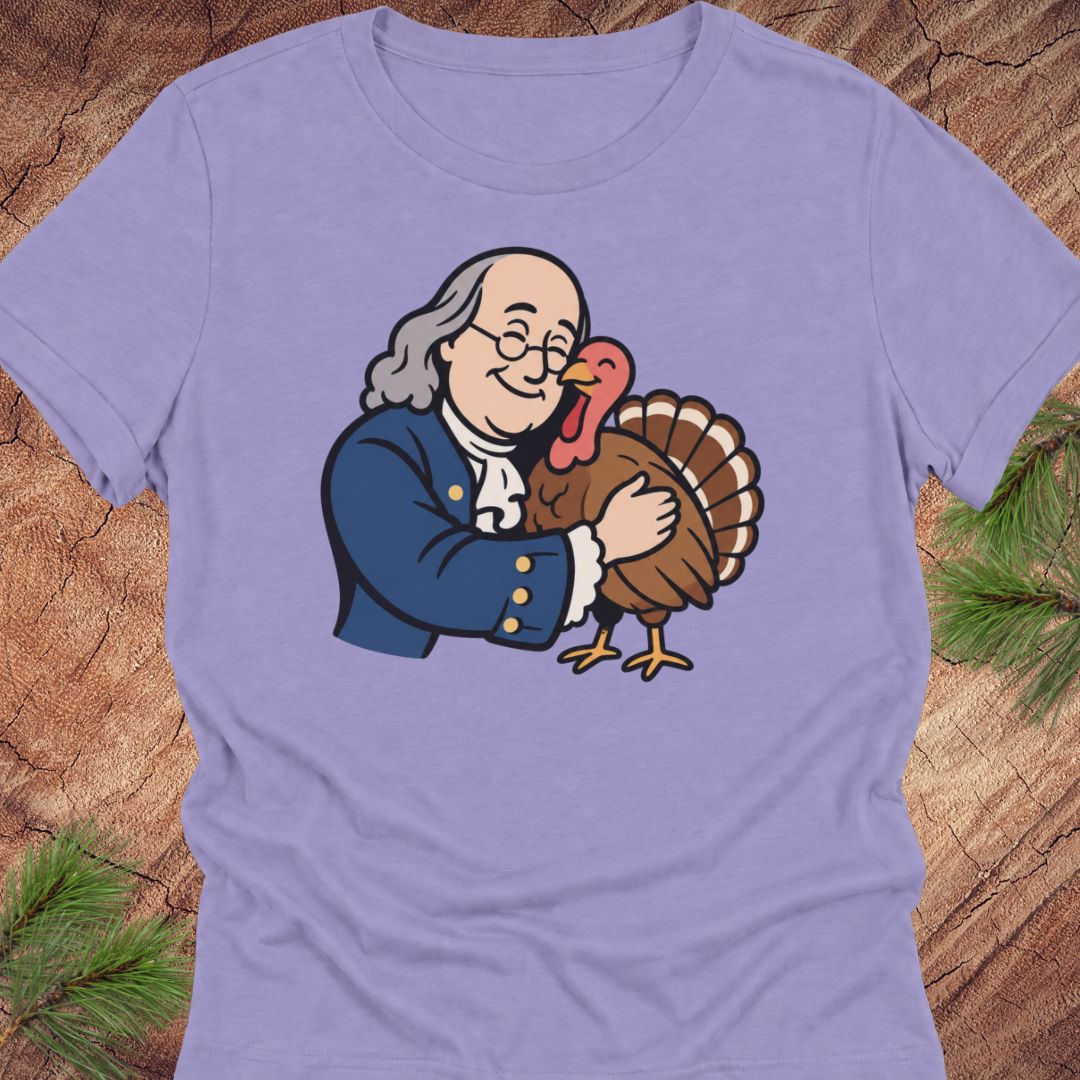 A light purple short-sleeve t-shirt with a cartoon graphic of Benjamin Franklin hugging a turkey, displayed flat on a rustic wood background with pine needles.