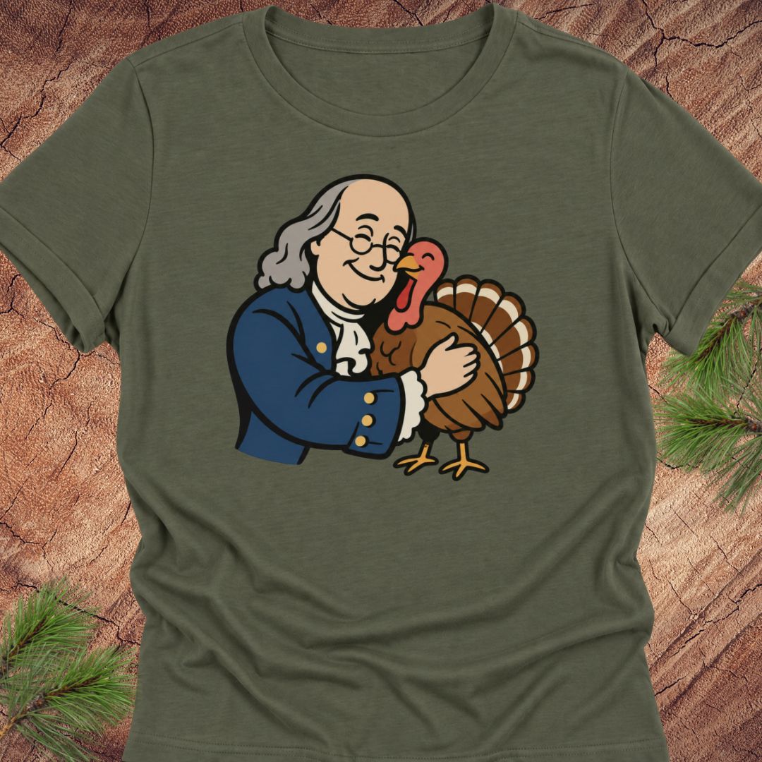 An olive green short-sleeve t-shirt featuring a cartoon graphic of Benjamin Franklin hugging a turkey, displayed flat on a rustic wood background with pine branches.