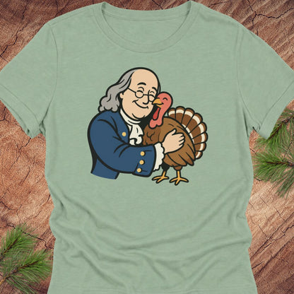 A sage green short-sleeve t-shirt featuring a cartoon graphic of George Washington hugging a turkey, displayed flat on a rustic wood background with pine needles.