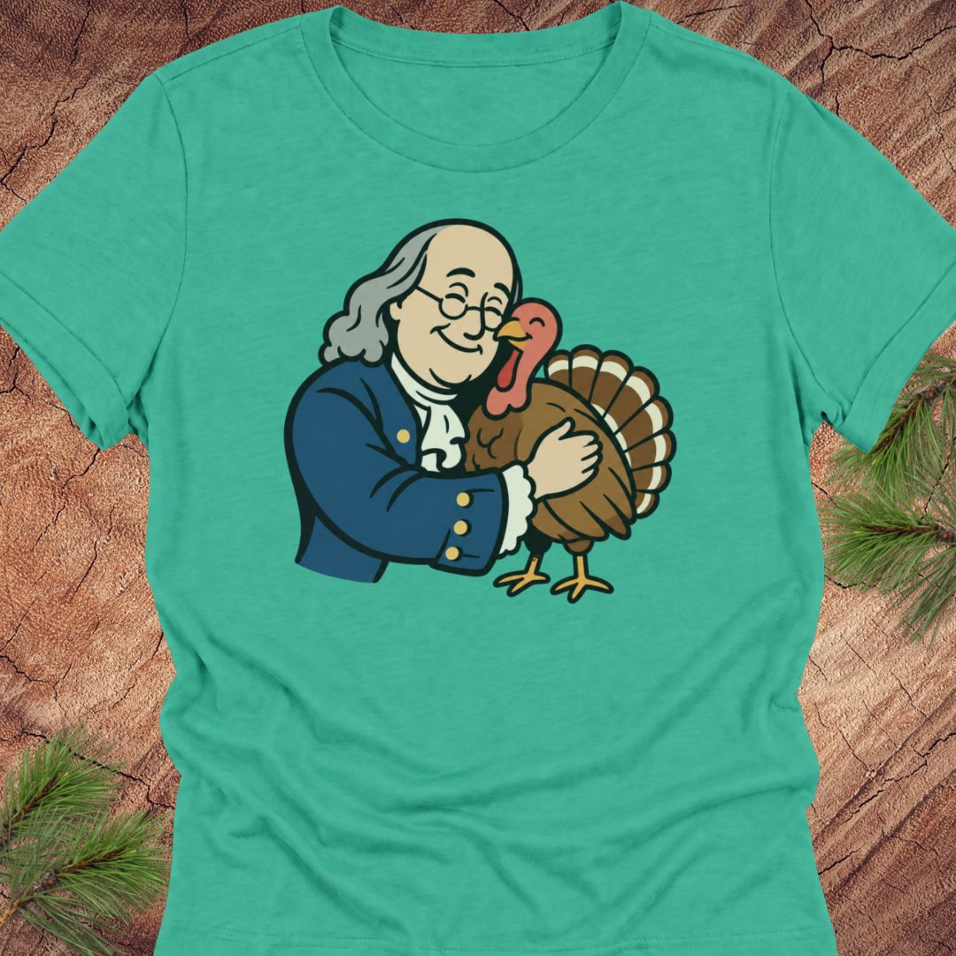 A green short-sleeve crewneck t-shirt featuring a cartoon graphic of Benjamin Franklin hugging a turkey, displayed flat on a rustic wood background with pine needles.