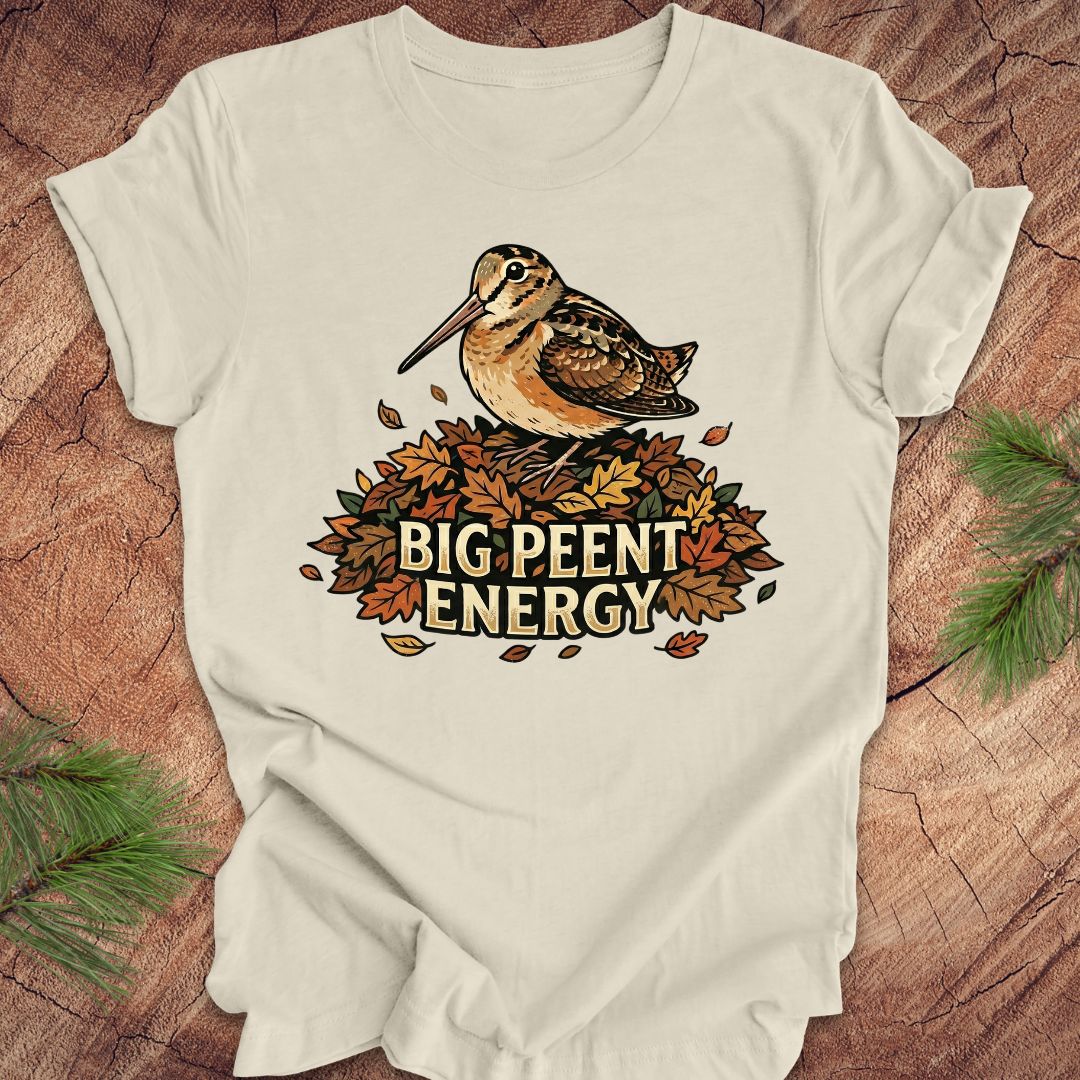 Beige t-shirt with a woodcock graphic and 'BIG PEENT ENERGY' text on a wooden surface.