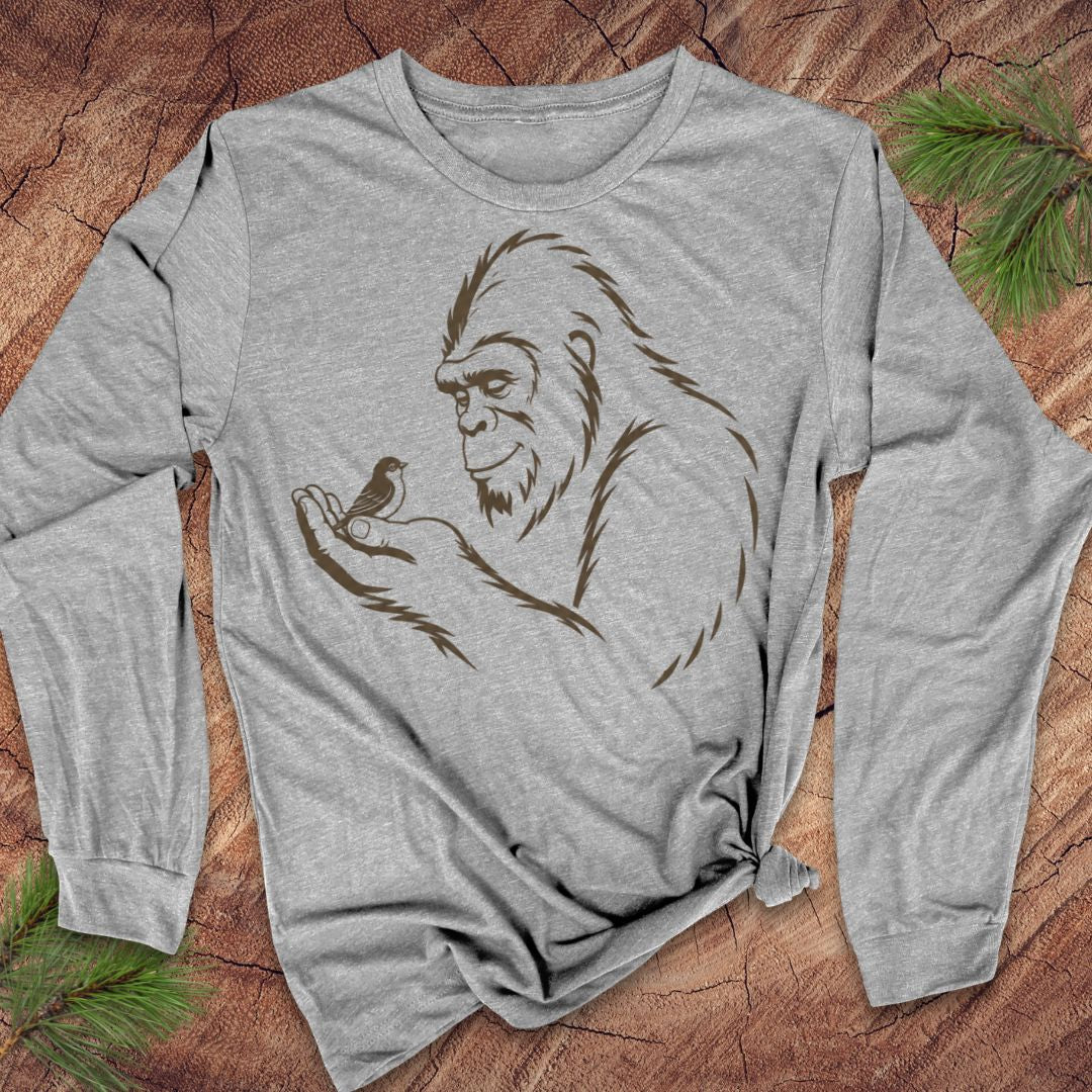 A heather gray long-sleeve t-shirt with a brown line art graphic of a Bigfoot holding a small bird, laid flat on a rustic wood background with pine branches.
