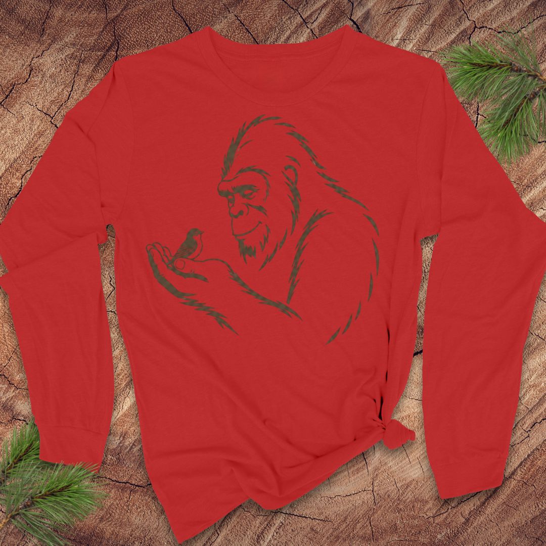 A red long-sleeve t-shirt with a dark green line art graphic of Bigfoot, styled flat on a rustic wood background with pine needles.