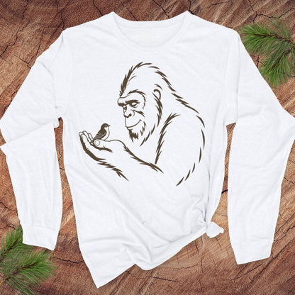 A white long-sleeve t-shirt with a dark brown line art graphic of bigfoot holding a small bird, laid flat on a rustic wood background with pine branches.