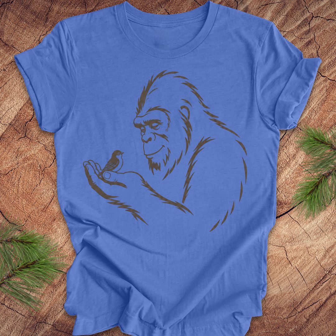 A blue short-sleeve crewneck t-shirt with a dark brown line art graphic of a Bigfoot figure holding a small bird, laid flat on a rustic wood background with pine needles in the corners.