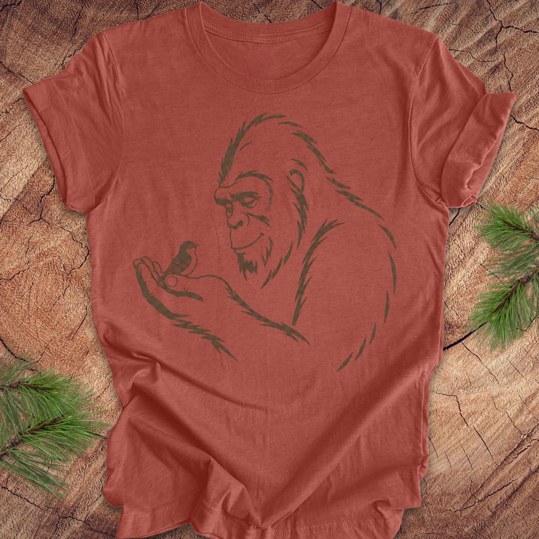 A terracotta-colored short-sleeve t-shirt with a dark green line art graphic of a Bigfoot holding a small bird, laid flat on a rustic wood slice background with pine needles in the corners.