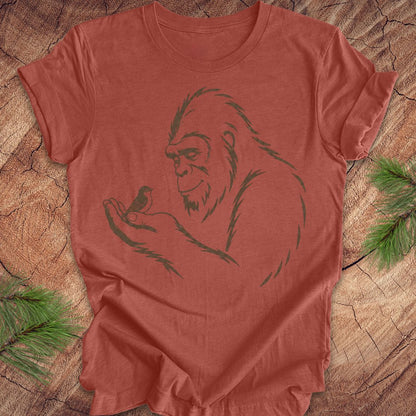 A terracotta-colored short-sleeve t-shirt with a dark green line art graphic of a Bigfoot holding a small bird, laid flat on a rustic wood slice background with pine needles in the corners.