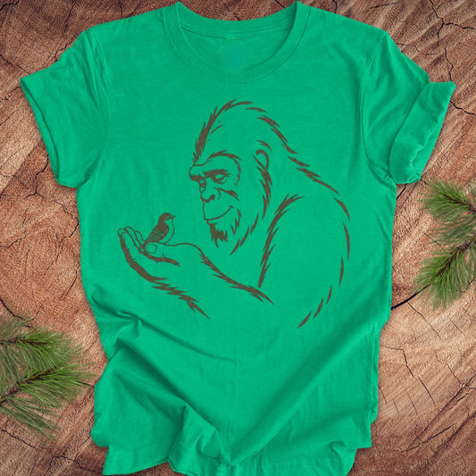 A bright green short-sleeve t-shirt with a dark brown graphic of a Bigfoot silhouette holding a small bird, laid flat on a rustic wood background with pine needles.