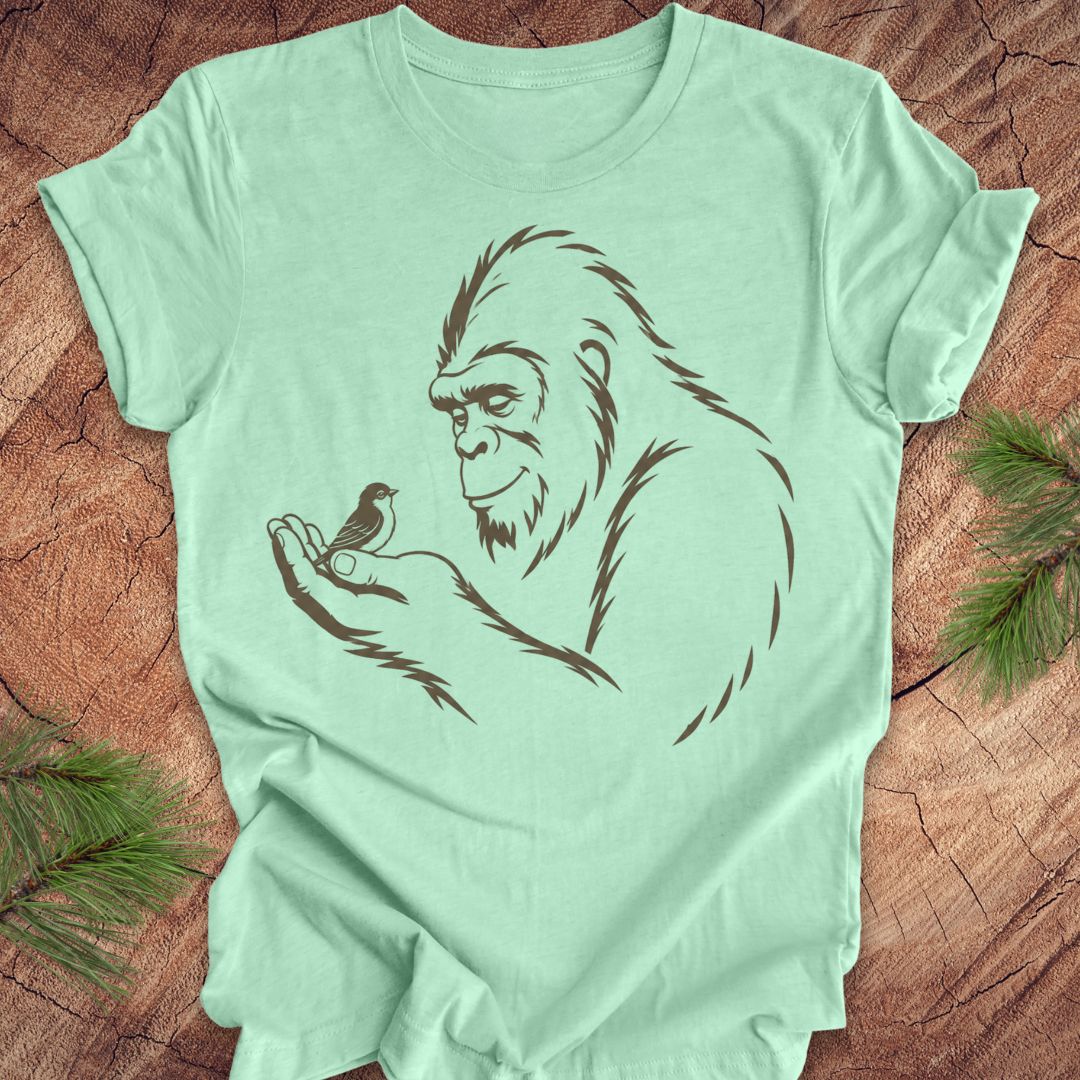 A mint green short-sleeve t-shirt with a dark brown graphic of a Bigfoot figure holding a small bird, laid flat on a rustic wood background with pine needles in the corners.