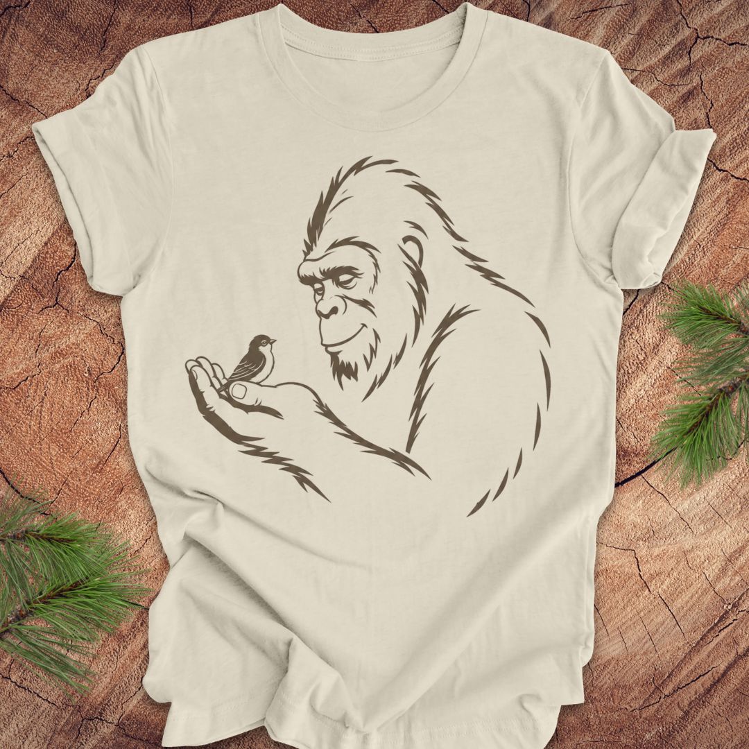 An beige short-sleeve t-shirt with a black line art graphic of a Bigfoot holding a small bird, laid flat on a rustic wood background with pine branches in the corners.