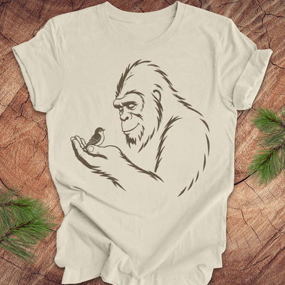 An beige short-sleeve t-shirt with a black line art graphic of a Bigfoot holding a small bird, laid flat on a rustic wood background with pine branches in the corners.