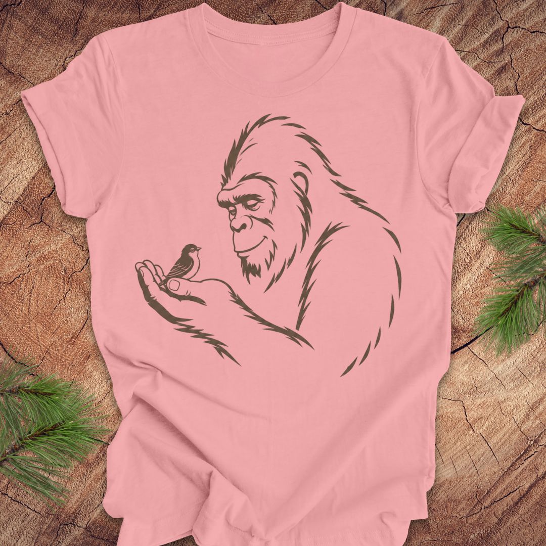A light pink short-sleeve t-shirt with a black line art graphic of a Bigfoot figure holding a small bird, laid flat on a rustic wood background with pine needles in the corners.