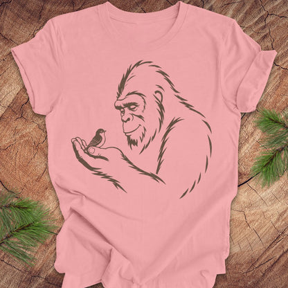 A light pink short-sleeve t-shirt with a black line art graphic of a Bigfoot figure holding a small bird, laid flat on a rustic wood background with pine needles in the corners.
