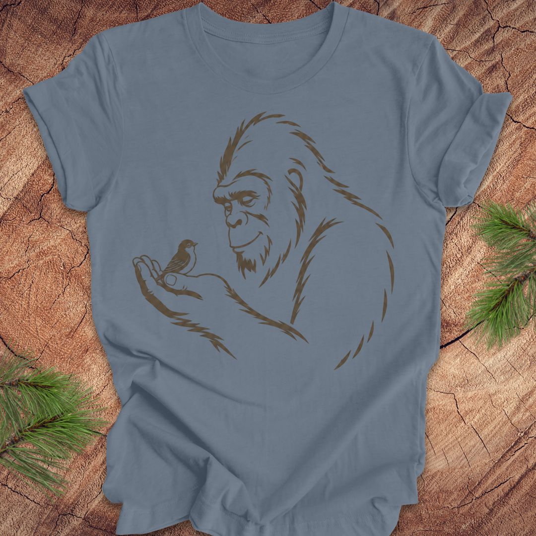 A dusty blue crewneck t-shirt featuring a brown line art graphic of a Bigfoot figure holding a small bird, laid flat on a rustic wood background with pine needles in the corners.