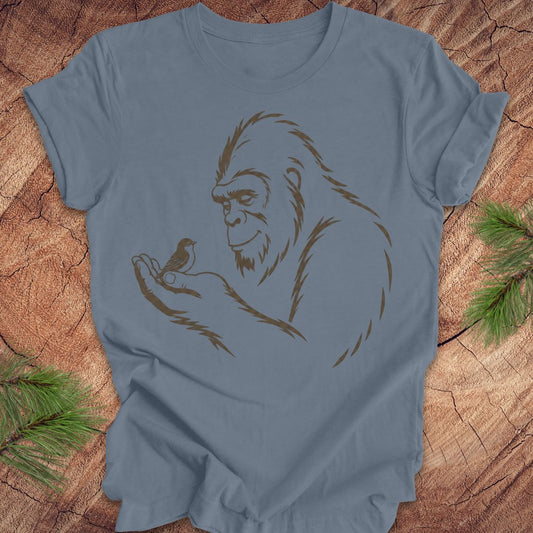 A dusty blue crewneck t-shirt featuring a brown line art graphic of a Bigfoot figure holding a small bird, laid flat on a rustic wood background with pine needles in the corners.