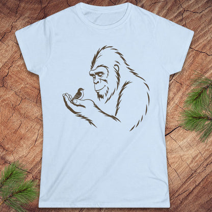A light blue short-sleeve t-shirt with a brown line art graphic of a Bigfoot holding a small bird, displayed flat on a rustic wood background with pine needles in the corners.