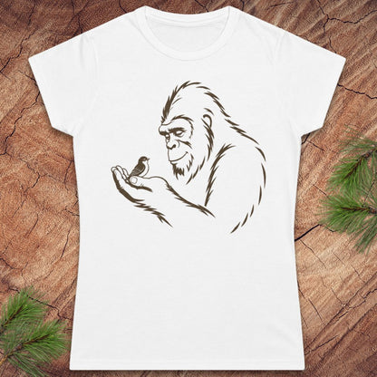 A white short-sleeve crewneck t-shirt featuring a brown line art graphic of a Bigfoot figure holding a small bird, displayed flat on a rustic wood background with pine branches in the corners.