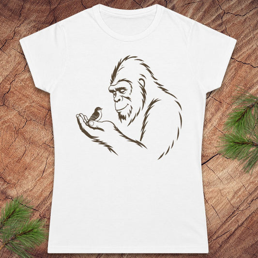 A white short-sleeve crewneck t-shirt featuring a brown line art graphic of a Bigfoot figure holding a small bird, displayed flat on a rustic wood background with pine branches in the corners.